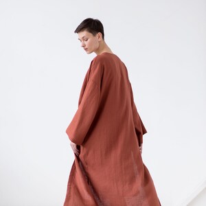 May include: A long, loose-fitting robe in a solid terracotta rust color. The garment features wide, three-quarter length sleeves and a simple, open-front design that falls to the mid-calf. The fabric has a visible, slightly textured weave.