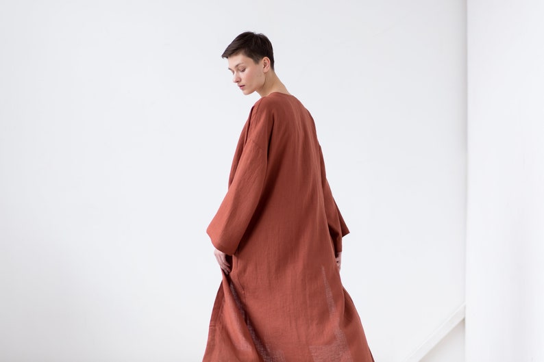 May include: A long, loose-fitting robe in a solid terracotta rust color. The garment features wide, three-quarter length sleeves and a simple, open-front design that falls to the mid-calf. The fabric has a visible, slightly textured weave.