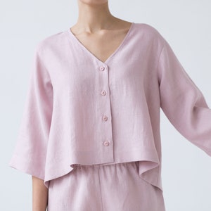 May include: A light pink linen top with a v-neck and button closure. The top has a cropped, boxy fit and 3/4 sleeves.