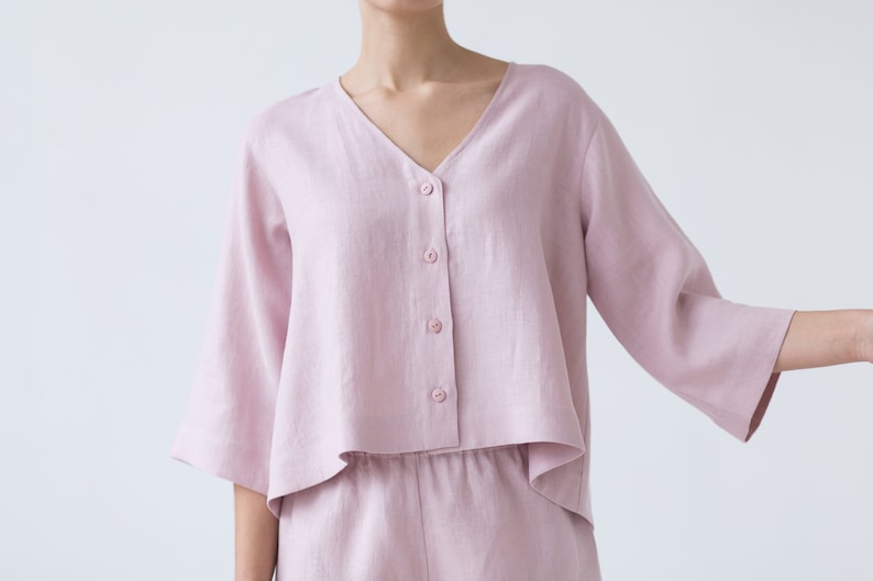 May include: A light pink linen top with a v-neck and button closure. The top has a cropped, boxy fit and 3/4 sleeves.