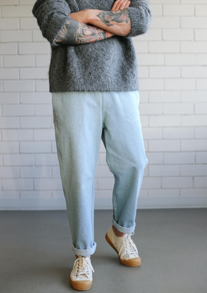 May include: A person wearing a gray sweater and light blue pants. The pants are cuffed at the ankles, paired with white sneakers with brown soles. The background is a white brick wall and a gray floor.