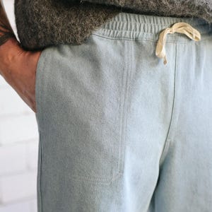 May include: Light blue pants with a drawstring waist and a pocket. The pants are made of a soft, textured fabric. A person's hand is in the pocket. The pants are part of a casual clothing set.
