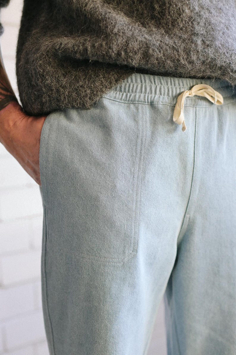 May include: Light blue pants with a drawstring waist and a pocket. The pants are made of a soft, textured fabric. A person's hand is in the pocket. The pants are part of a casual clothing set.