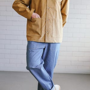 Puede incluir: Una persona vestida con una chaqueta abotonada color beige con dos bolsillos delanteros y vaqueros azul claro. La persona tambi&eacute;n lleva zapatillas blancas con suelas beige. El atuendo es informal y elegante.