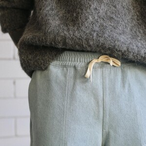 May include: A pair of light blue sweatpants featuring an elastic waistband with a cream-colored drawstring, paired with a dark gray fuzzy sweater. The sweatpants have a visible vertical seam down the front center of each leg.