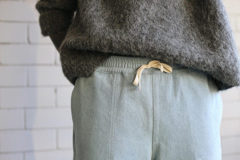 May include: A pair of light blue sweatpants featuring an elastic waistband with a cream-colored drawstring, paired with a dark gray fuzzy sweater. The sweatpants have a visible vertical seam down the front center of each leg.