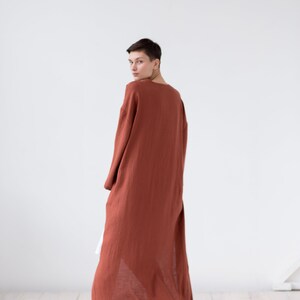 May include: A long-sleeved, floor-length robe in a solid terracotta rust color. The garment features a loose, flowing silhouette with a simple round neckline and wide sleeves, crafted from a lightweight, textured fabric that drapes to the floor.