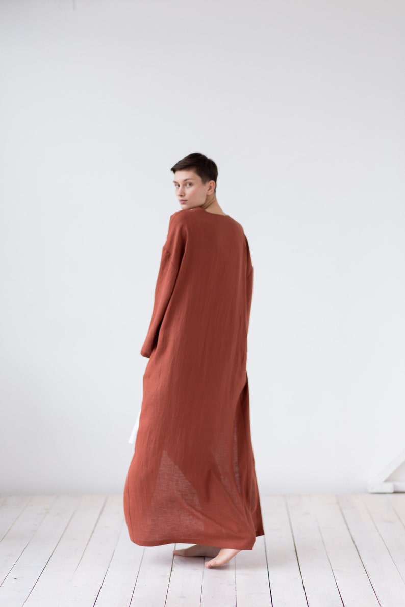 May include: A long-sleeved, floor-length robe in a solid terracotta rust color. The garment features a loose, flowing silhouette with a simple round neckline and wide sleeves, crafted from a lightweight, textured fabric that drapes to the floor.