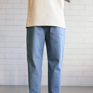 Puede incluir: Pantalones vaqueros azul claro con dobladillos vueltos. Los pantalones tienen un bolsillo trasero y se combinan con una camiseta color crema y zapatos de lona blancos. El fondo es una pared de ladrillos blancos.