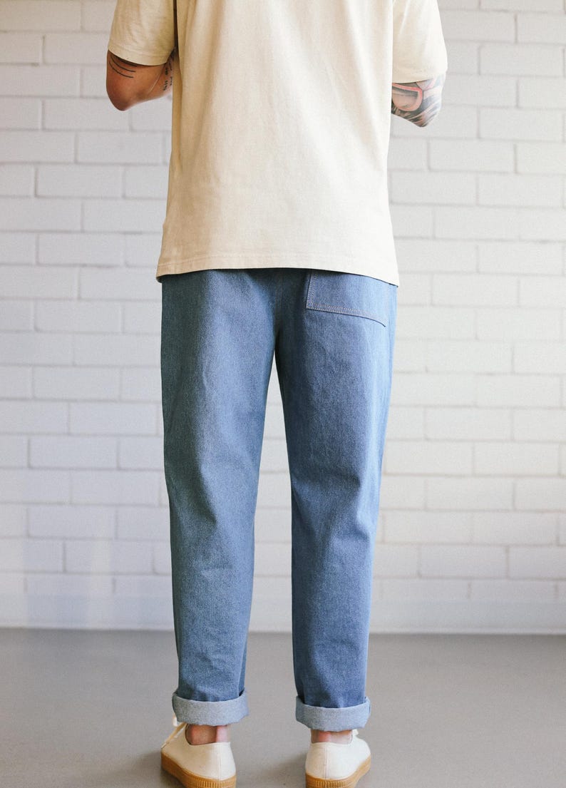Puede incluir: Pantalones vaqueros azul claro con dobladillos vueltos. Los pantalones tienen un bolsillo trasero y se combinan con una camiseta color crema y zapatos de lona blancos. El fondo es una pared de ladrillos blancos.