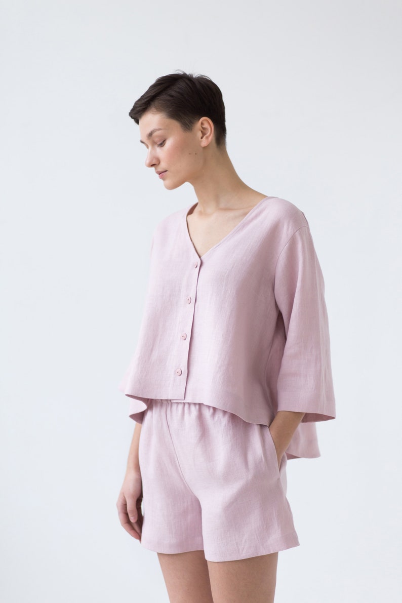 May include: A light pink linen button-up shirt with a v-neck and short sleeves, paired with matching linen shorts. The shorts have a drawstring waist and pockets.