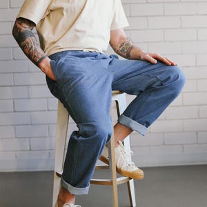 Puede incluir: Una persona con camiseta crema y vaqueros azules con dobladillos, sentada en un taburete blanco. Lleva zapatillas blancas con suela color canela. El fondo es una pared de ladrillos blancos.