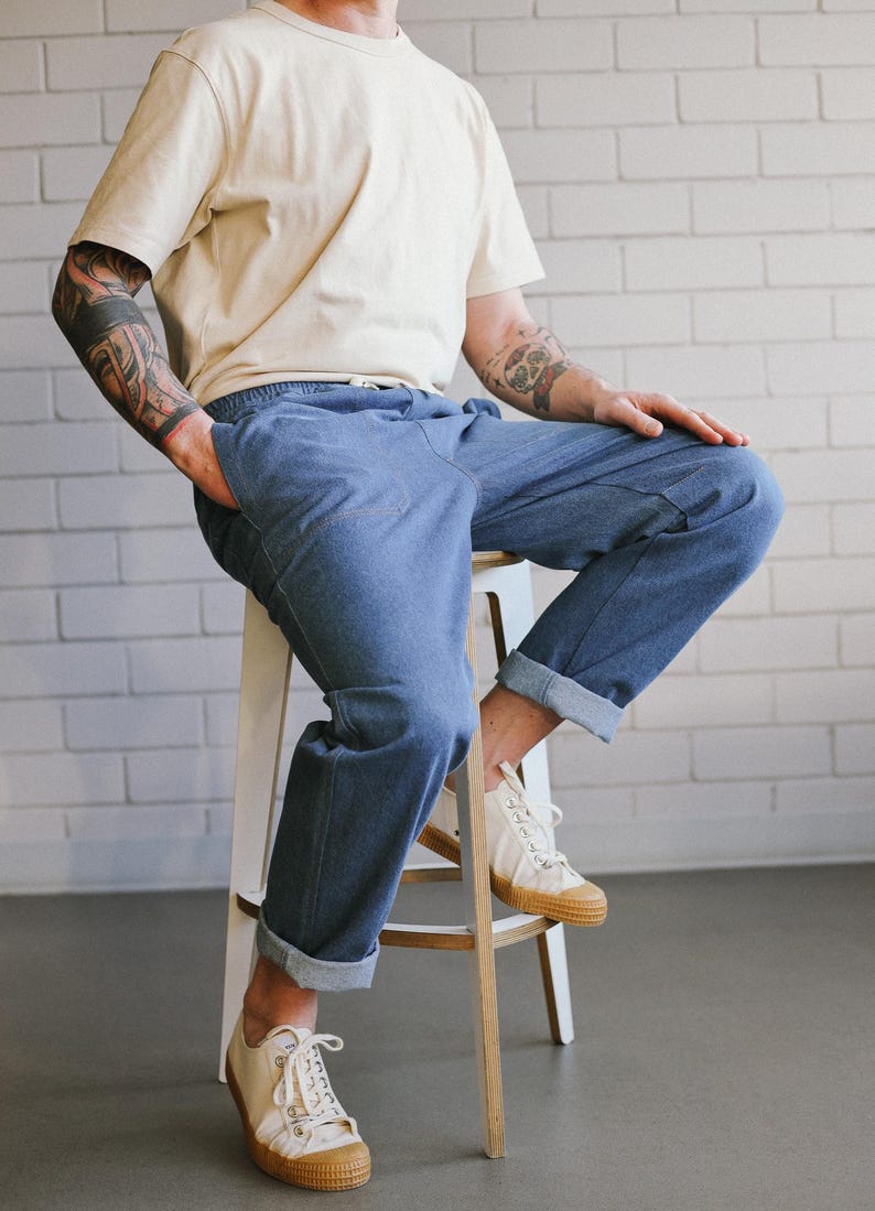 Puede incluir: Una persona con camiseta crema y vaqueros azules con dobladillos, sentada en un taburete blanco. Lleva zapatillas blancas con suela color canela. El fondo es una pared de ladrillos blancos.