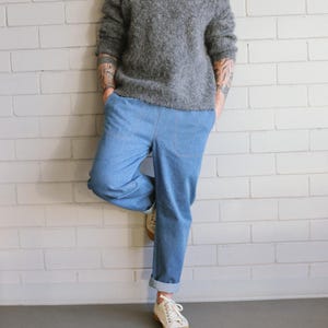 Puede incluir: Una persona con un su&eacute;ter gris y borroso y pantalones vaqueros azul claro con los pu&ntilde;os doblados. La persona lleva zapatillas blancas con suela color canela. La persona est&aacute; apoyada en una pared de ladrillos blancos.