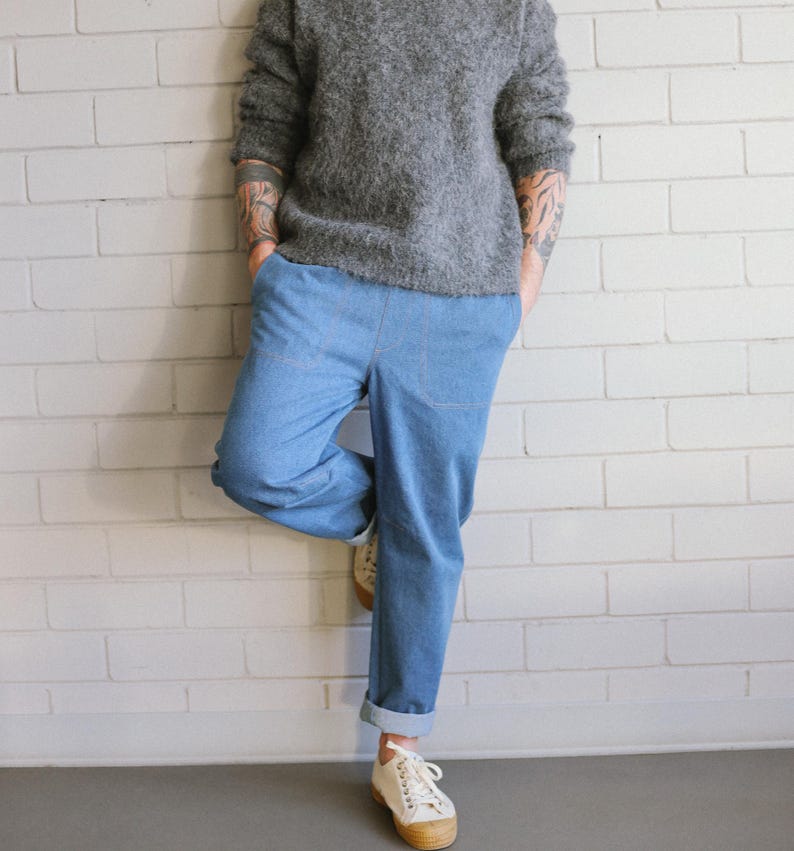 Puede incluir: Una persona con un su&eacute;ter gris y borroso y pantalones vaqueros azul claro con los pu&ntilde;os doblados. La persona lleva zapatillas blancas con suela color canela. La persona est&aacute; apoyada en una pared de ladrillos blancos.