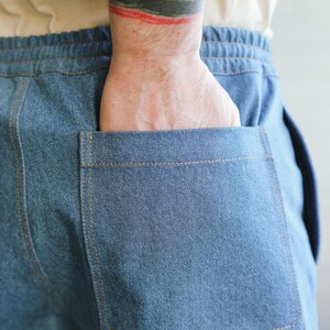 Puede incluir: Primer plano de pantalones vaqueros azules con cintura con cord&oacute;n y un bolsillo trasero grande. El bolsillo est&aacute; cosido con hilo color beige. Una mano est&aacute; en el bolsillo.