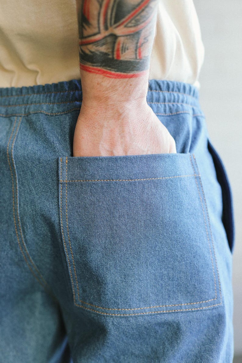 Puede incluir: Primer plano de pantalones vaqueros azules con cintura con cord&oacute;n y un bolsillo trasero grande. El bolsillo est&aacute; cosido con hilo color beige. Una mano est&aacute; en el bolsillo.