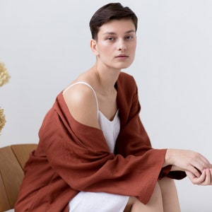 May include: A white camisole with thin straps worn under a loose-fitting, long-sleeved, rust-colored robe. The robe features wide sleeves and a textured fabric finish, draped over the shoulders to reveal the white garment underneath.