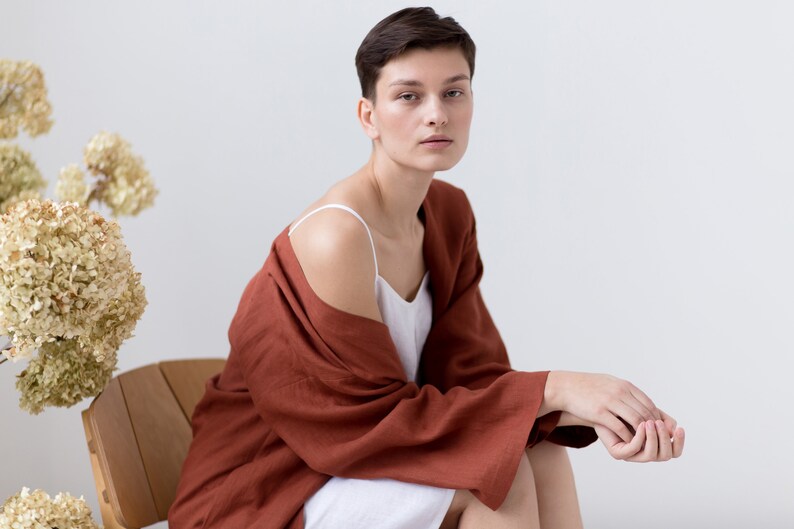 May include: A white camisole with thin straps worn under a loose-fitting, long-sleeved, rust-colored robe. The robe features wide sleeves and a textured fabric finish, draped over the shoulders to reveal the white garment underneath.