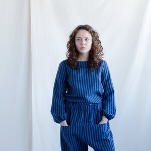 May include: A long-sleeved top and matching pants set featuring a vertical striped pattern in two shades of blue. The top has a round neckline and the pants include an elasticized waistband with two large front pockets and gathered cuffs.