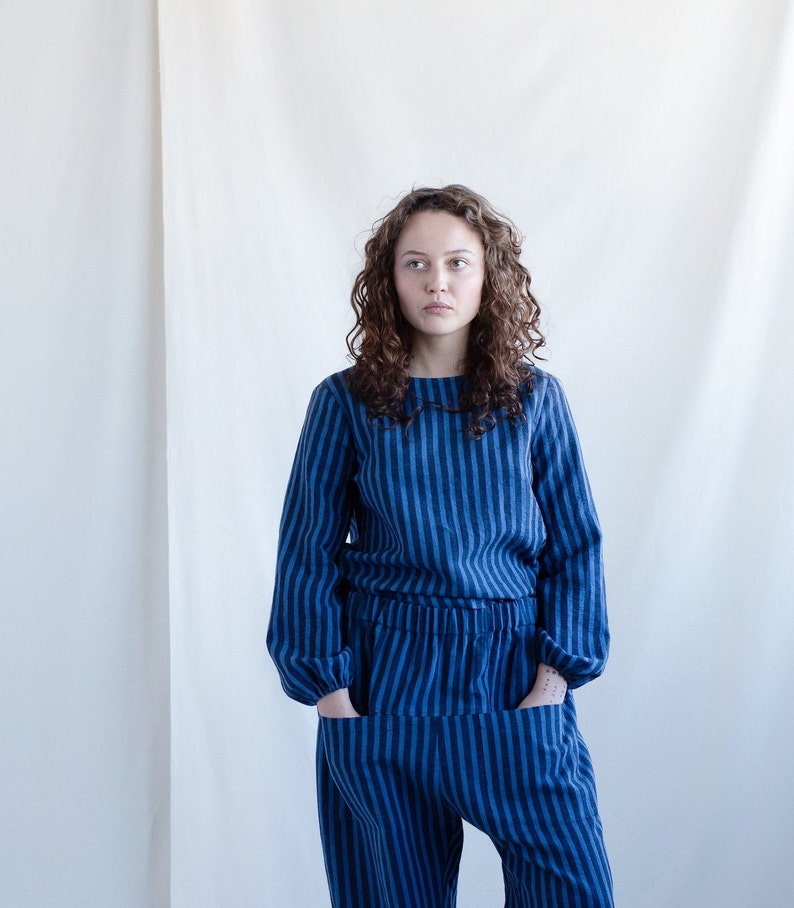 May include: A long-sleeved top and matching pants set featuring a vertical striped pattern in two shades of blue. The top has a round neckline and the pants include an elasticized waistband with two large front pockets and gathered cuffs.