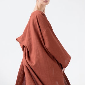 May include: A loose-fitting, long-sleeved garment in a solid terracotta orange color. The piece features a wide, draped neckline and a relaxed, flowing silhouette with visible fabric texture, extending down to cover the torso and upper legs.