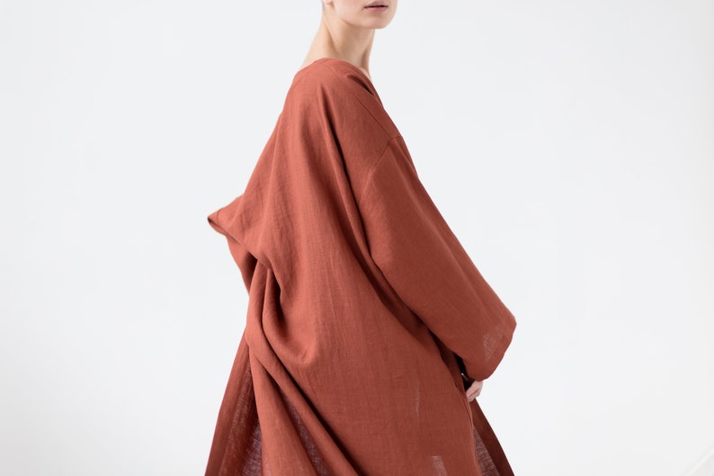 May include: A loose-fitting, long-sleeved garment in a solid terracotta orange color. The piece features a wide, draped neckline and a relaxed, flowing silhouette with visible fabric texture, extending down to cover the torso and upper legs.