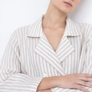 May include: A double-breasted blazer featuring a vertical striped pattern in beige and white. The garment has a notched lapel collar, long sleeves, and a single visible white button closure at the front, crafted from a textured fabric.