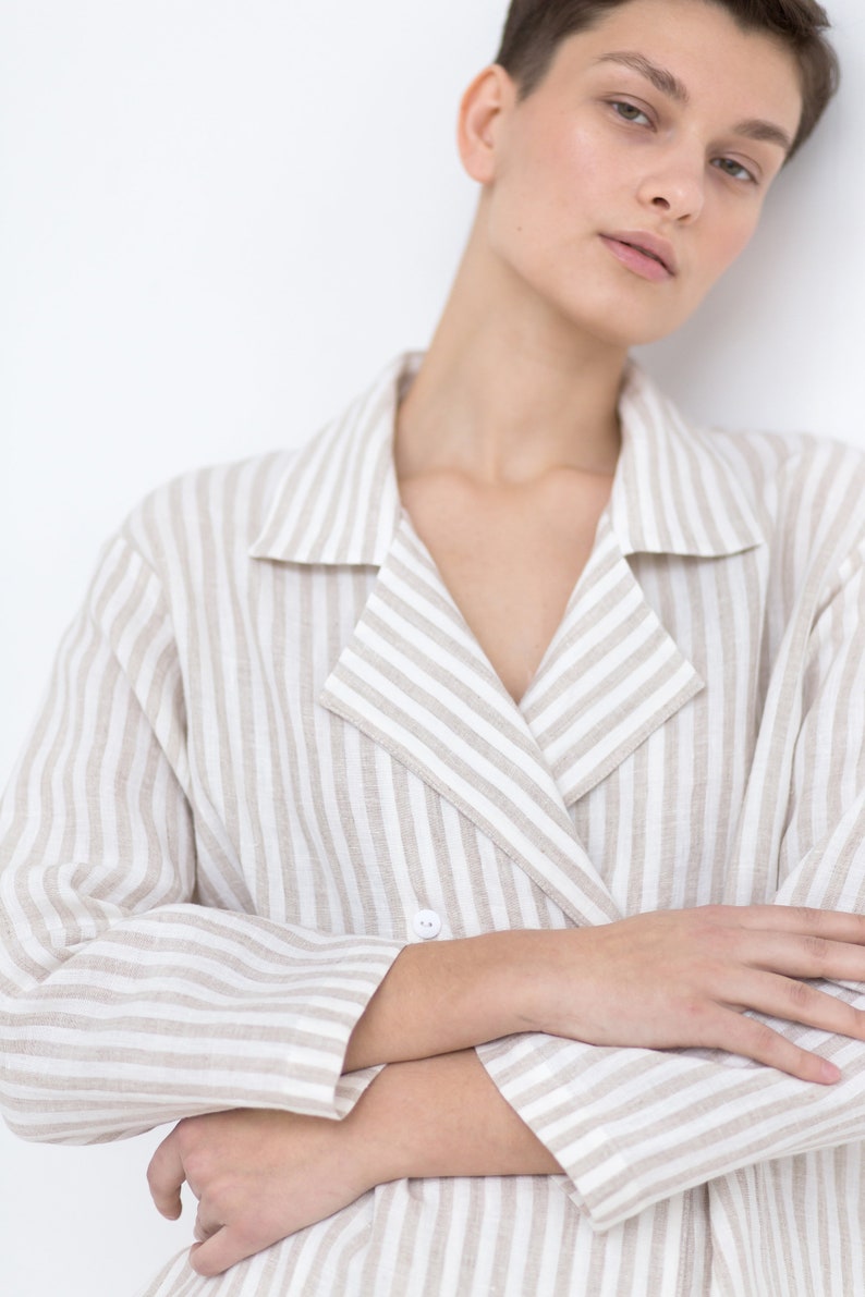 May include: A double-breasted blazer featuring a vertical striped pattern in beige and white. The garment has a notched lapel collar, long sleeves, and a single visible white button closure at the front, crafted from a textured fabric.