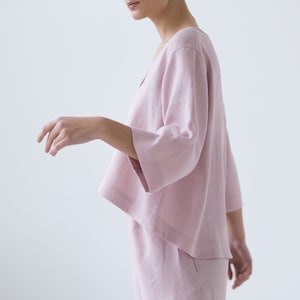 May include: A pale pink linen top with a relaxed fit and three-quarter sleeves. The top has a V-neckline and a slightly cropped hem.
