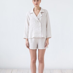 May include: A two-piece pajama set featuring a double-breasted long-sleeve top with a notched collar and four white buttons, paired with matching shorts. The garment is made of a light beige and white vertical striped fabric with a textured finish.