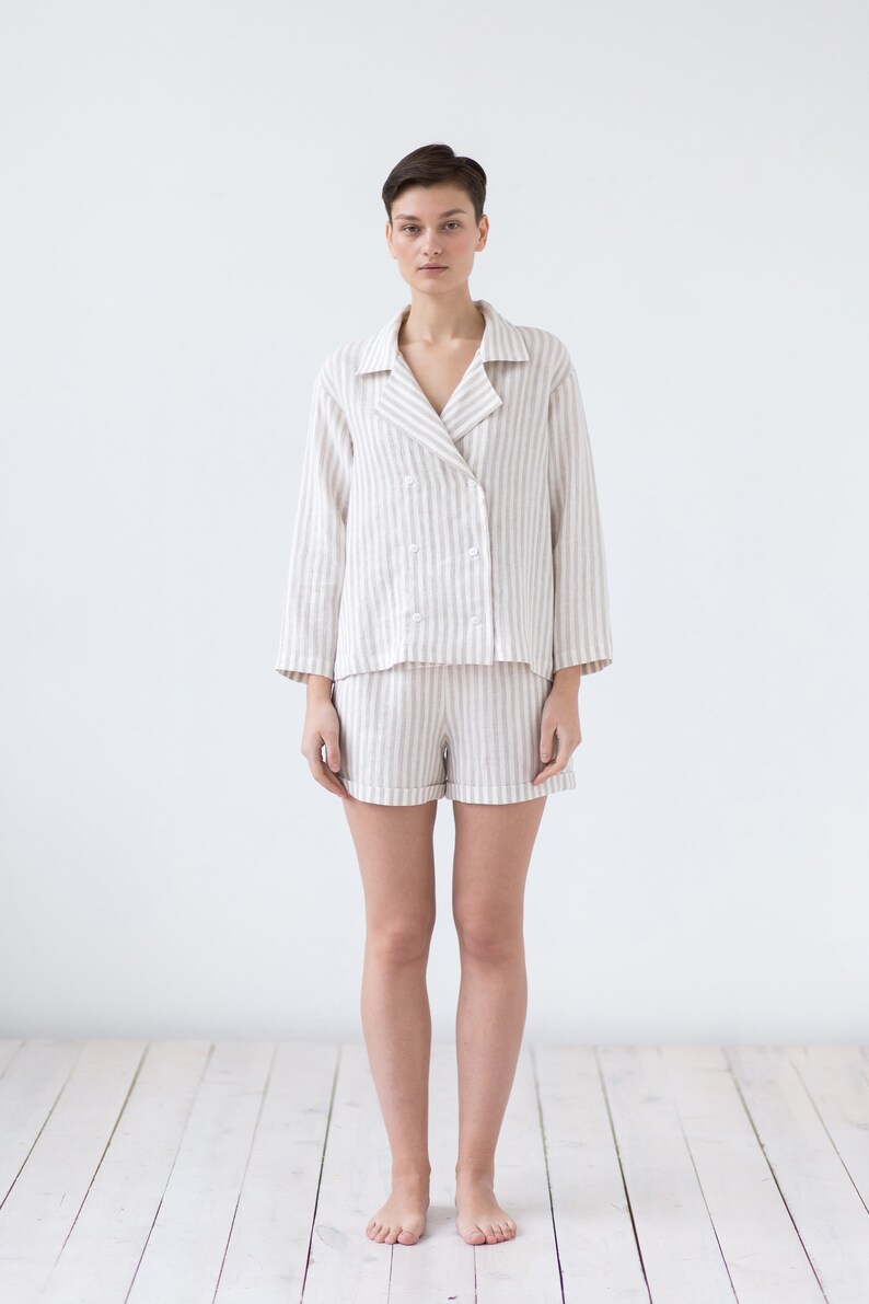 May include: A two-piece pajama set featuring a double-breasted long-sleeve top with a notched collar and four white buttons, paired with matching shorts. The garment is made of a light beige and white vertical striped fabric with a textured finish.
