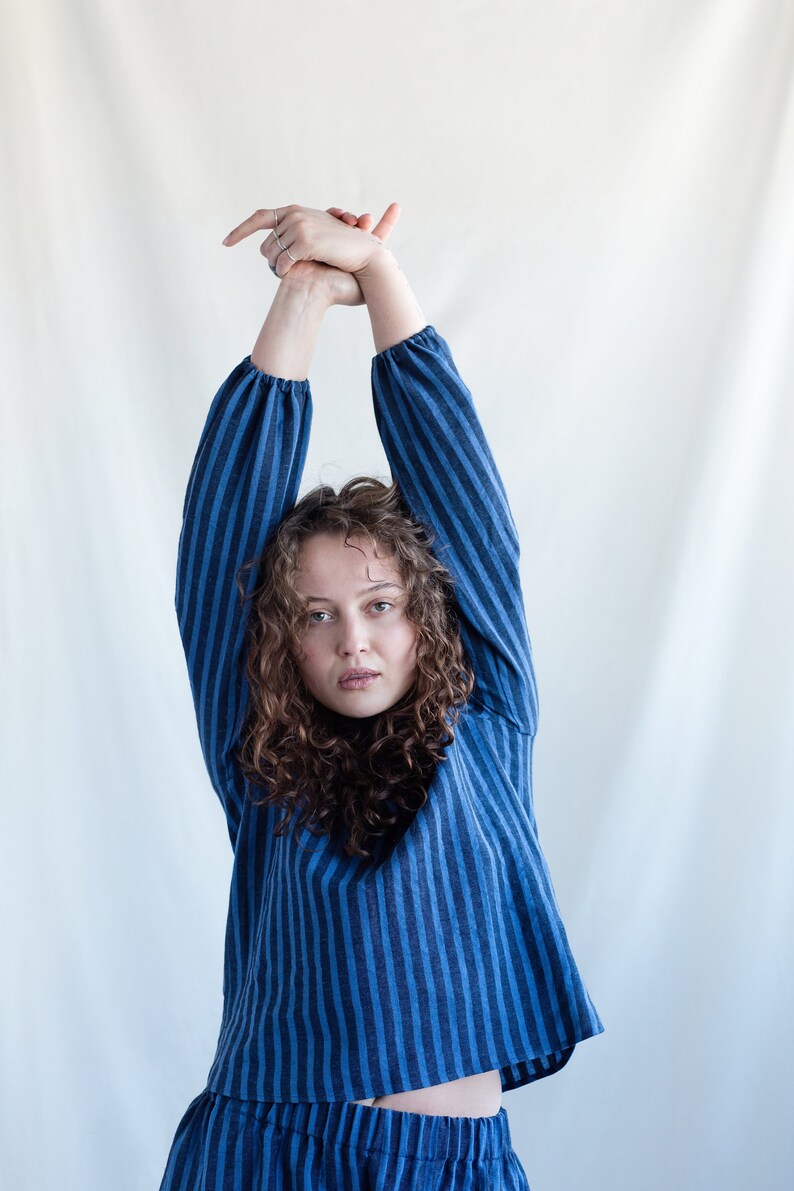 May include: A long-sleeved top featuring a vertical striped pattern in shades of dark blue and lighter blue. The garment has a relaxed fit, a simple round neckline, and gathered cuffs at the wrists, creating a soft, draped silhouette in a woven fabric.