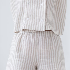 May include: A two-piece set consisting of a double-breasted short-sleeve shirt and matching shorts. Both pieces feature a vertical striped pattern in beige and white. The shirt has a notched collar and four white buttons on the front closure.