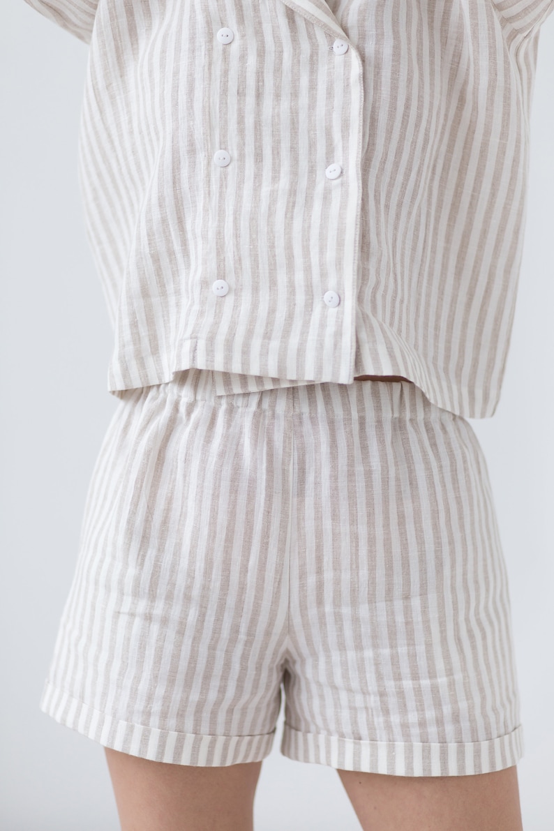 May include: A two-piece set consisting of a double-breasted short-sleeve shirt and matching shorts. Both pieces feature a vertical striped pattern in beige and white. The shirt has a notched collar and four white buttons on the front closure.