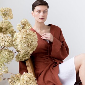 May include: A long-sleeved, wrap-style robe in a solid rust-brown color, featuring a V-neckline and a matching fabric tie belt at the waist. The garment is worn over a simple white undergarment, showcasing a clean, structured design.