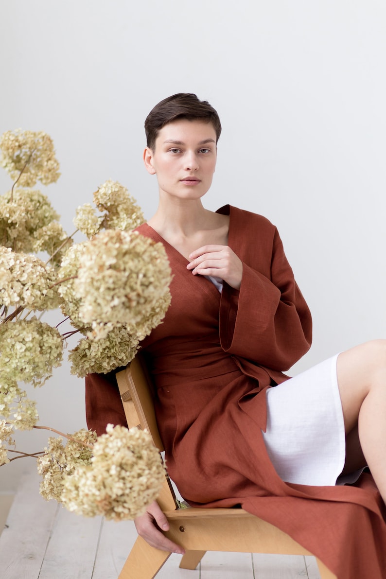 May include: A long-sleeved, wrap-style robe in a solid rust-brown color, featuring a V-neckline and a matching fabric tie belt at the waist. The garment is worn over a simple white undergarment, showcasing a clean, structured design.