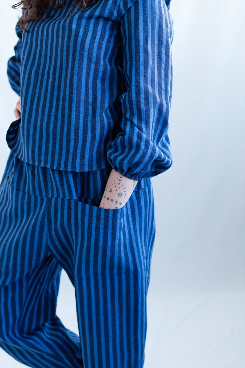 May include: A long-sleeved top and matching pants set featuring a vertical striped pattern in alternating shades of dark blue and bright blue. The top has gathered cuffs at the wrists, and the pants include a visible pocket on the front side.