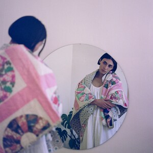 May include: A person stands in front of a mirror wearing a white dress and a colorful patchwork quilt. The quilt has pink, green, and white fabric with floral patterns.