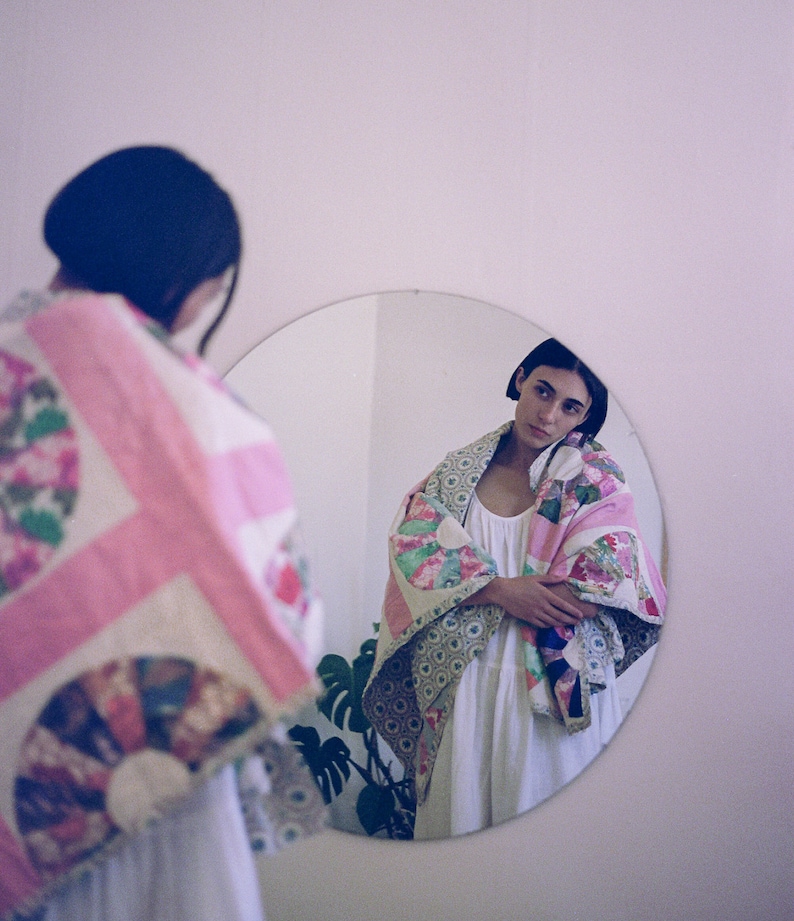 May include: A person stands in front of a mirror wearing a white dress and a colorful patchwork quilt. The quilt has pink, green, and white fabric with floral patterns.