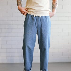 Puede incluir: Pantalones vaqueros azul claro con cintura con cord&oacute;n y pu&ntilde;os doblados. Los pantalones tienen dos bolsillos delanteros y se combinan con una camiseta color crema y zapatillas blancas. El fondo es una pared de ladrillos blancos.
