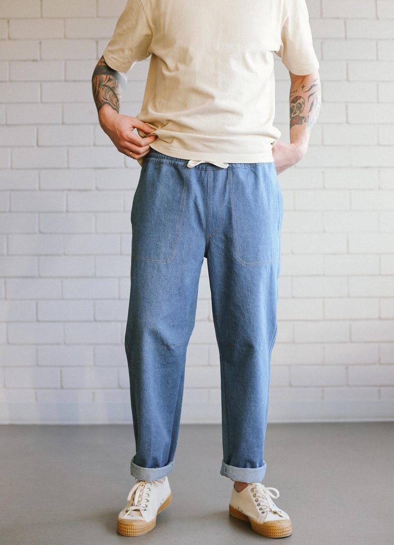 Puede incluir: Pantalones vaqueros azul claro con cintura con cord&oacute;n y pu&ntilde;os doblados. Los pantalones tienen dos bolsillos delanteros y se combinan con una camiseta color crema y zapatillas blancas. El fondo es una pared de ladrillos blancos.