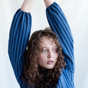 May include: A long-sleeved shirt featuring a vertical striped pattern in alternating shades of dark blue and light blue. The garment has gathered cuffs at the wrists and a simple neckline, constructed from a textured fabric with a visible weave.