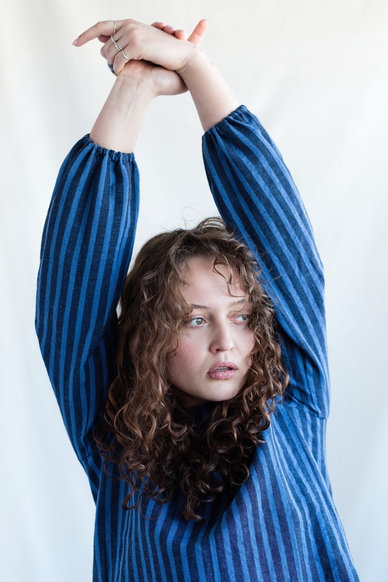 May include: A long-sleeved shirt featuring a vertical striped pattern in alternating shades of dark blue and light blue. The garment has gathered cuffs at the wrists and a simple neckline, constructed from a textured fabric with a visible weave.