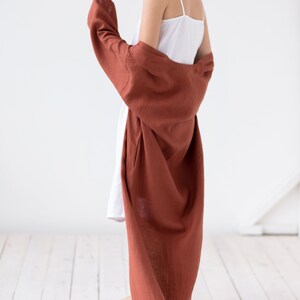 May include: A white camisole dress with thin spaghetti straps paired with a long, loose-fitting, rust-colored robe. The robe features long sleeves and a textured fabric finish, draping down to the floor while worn over the white dress.