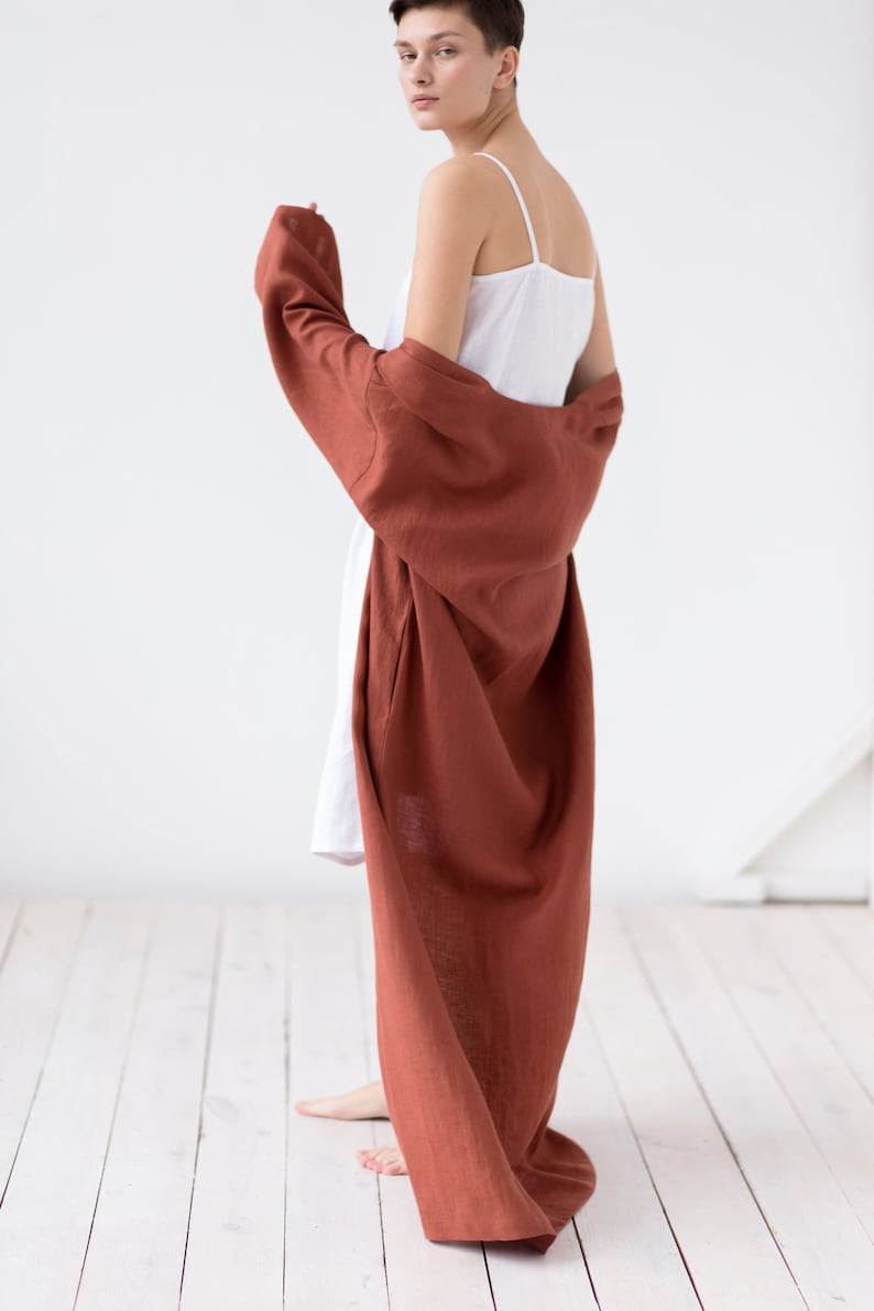 May include: A white camisole dress with thin spaghetti straps paired with a long, loose-fitting, rust-colored robe. The robe features long sleeves and a textured fabric finish, draping down to the floor while worn over the white dress.