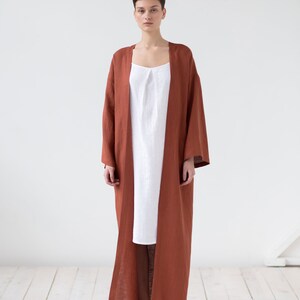 May include: A long, open-front rust-colored robe with wide sleeves layered over a simple white sleeveless dress. The robe features a straight, floor-length silhouette and a plain texture, while the white dress has a subtle pleated neckline.