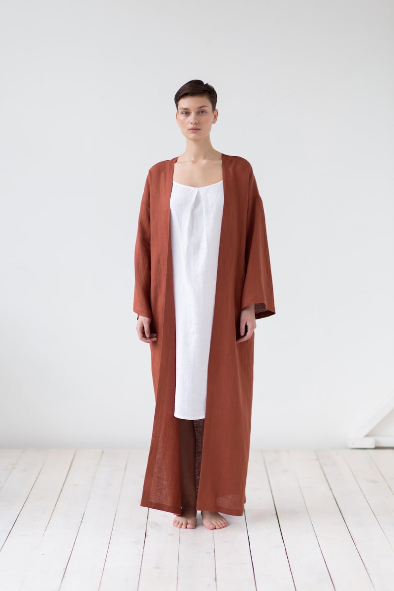 May include: A long, open-front rust-colored robe with wide sleeves layered over a simple white sleeveless dress. The robe features a straight, floor-length silhouette and a plain texture, while the white dress has a subtle pleated neckline.