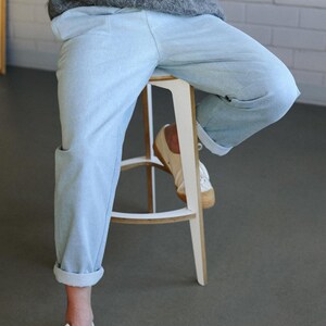 May include: Light blue denim pants with rolled cuffs. The pants are paired with white canvas sneakers with a brown sole. The person is wearing a gray sweater and sitting on a white stool with a light brown seat.