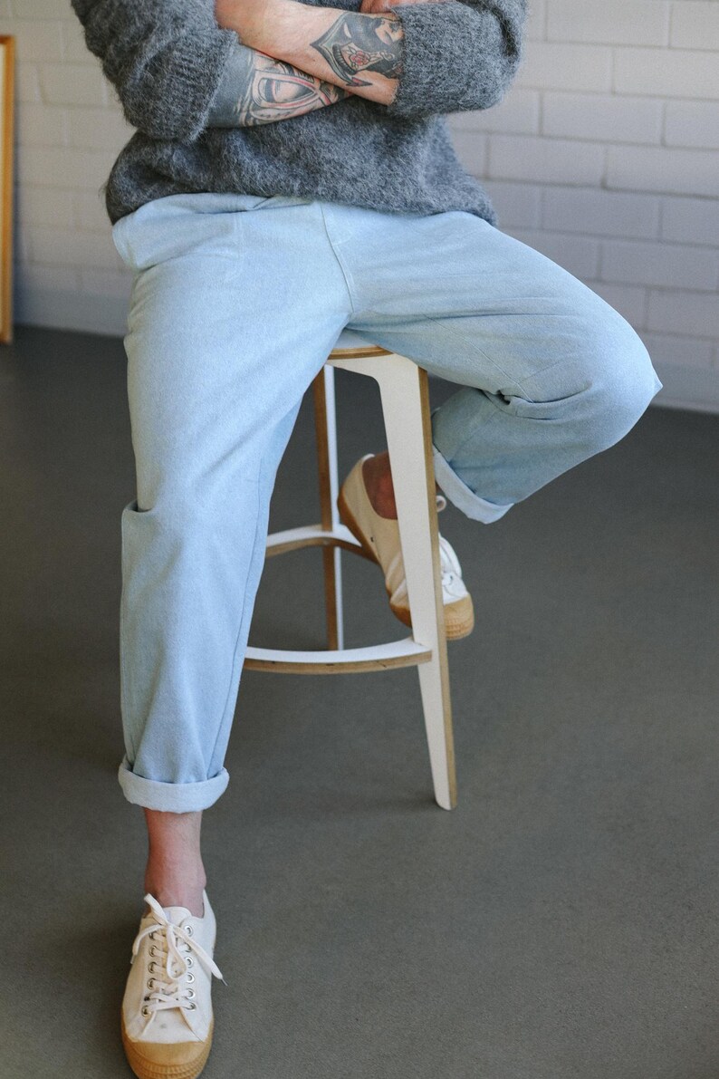 May include: Light blue denim pants with rolled cuffs. The pants are paired with white canvas sneakers with a brown sole. The person is wearing a gray sweater and sitting on a white stool with a light brown seat.